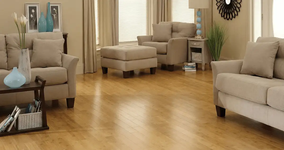 A living room with professionally installed bamboo flooring, beige sofas, a matching ottoman, a side table with a blue lamp, vases, framed pictures, and large windows with blinds and curtains in Tallahassee, FL